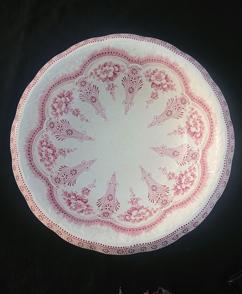 May include: A round, white ceramic serving platter with a scalloped edge. The platter features a pink floral and geometric design around the rim and center. The floral pattern includes detailed flowers and decorative elements.