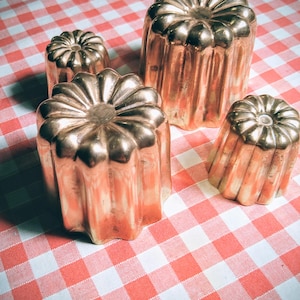 Peut inclure: Quatre moules à cannelés en cuivre de différentes tailles sont disposés sur une nappe à carreaux rouges et blancs. Les moules ont une forme cylindrique cannelée avec un design supérieur distinctif. Le cuivre a un éclat chaud et réfléchissant.