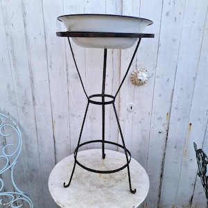 May include: A white enamel basin with a dark blue rim resting on a tall, black, three-legged metal stand. The stand features a circular support ring in the middle and curved feet at the base, providing a stable structure for the basin.
