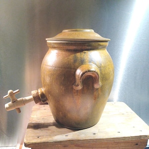 May include: A large, round, mustard-yellow ceramic crock with a matching lid and two side handles. A light-coloured wooden spigot with a cork stopper is inserted into the lower front section of the vessel, allowing for liquid dispensing.