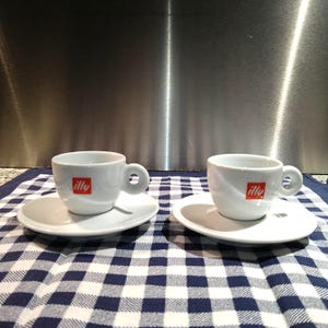 Könnte beinhalten: Zwei weiße Espressotassen und Untertassen mit dem roten "illy"-Logo. Die Tassen haben einen runden Henkel und stehen auf kleinen, runden Untertassen. Das Set steht auf einem blau-weiß karierten Tuch.