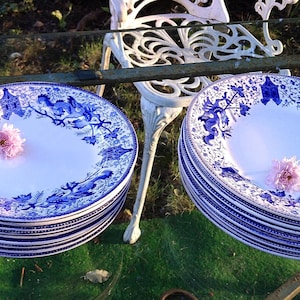 May include: A stack of white ceramic plates with a blue floral design and a pink flower in the center. The plates are stacked on top of each other. The plates are outdoors, with a white chair in the background.