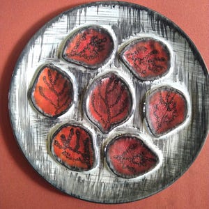 May include: A ceramic plate with a unique design. The plate features seven leaf-shaped indentations, each filled with a red and black leaf pattern. The plate's rim has a black and white brushstroke design.