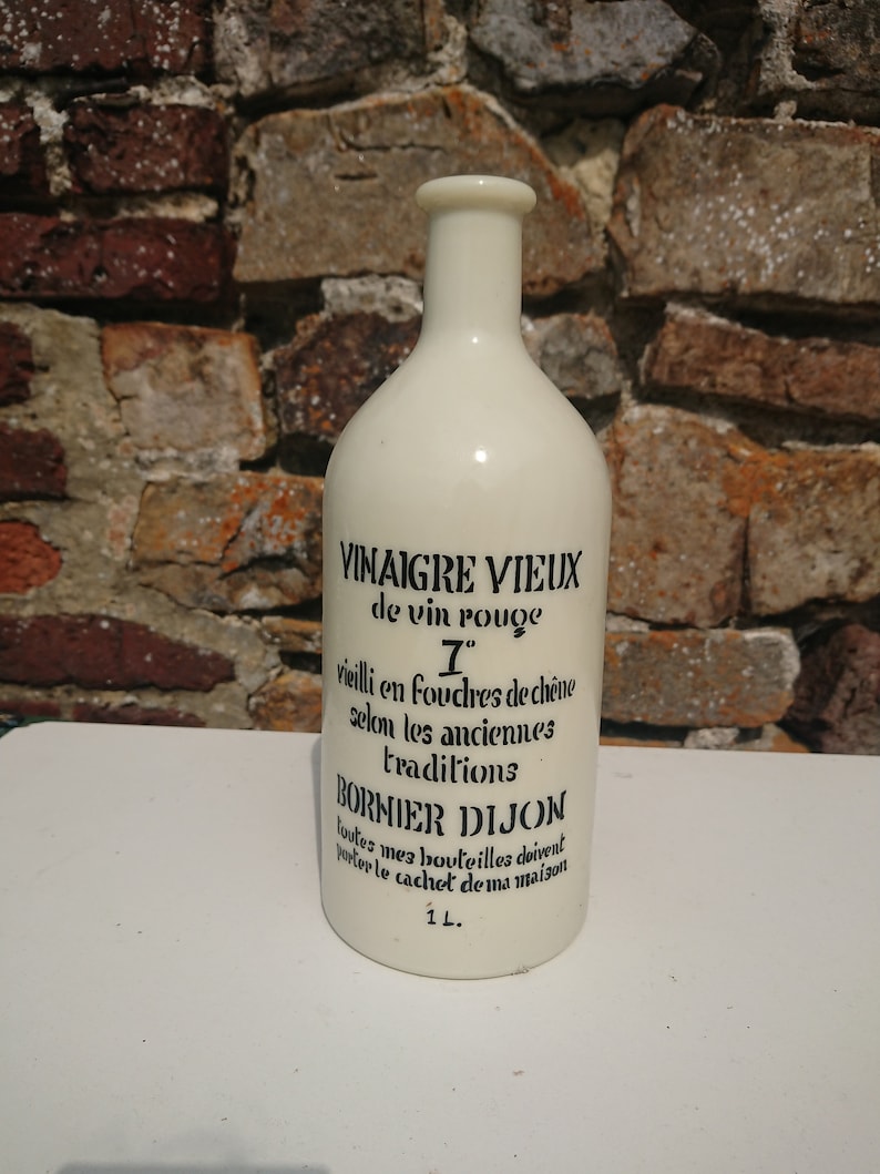 Puede incluir: Botella de cer&aacute;mica blanquecina con texto negro. La botella dice "VINAIGRE VIEUX de vin rouge" y "BORNIER DIJON". La botella tiene un volumen de 1 litro. El fondo es una pared de ladrillos.