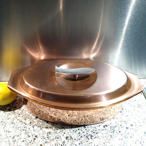 以下が含まれることがあります： 蓋付きの楕円形銅製キャセロール皿。蓋には魚の形をした銀色の金属製ハンドルが付いています。皿の外側は磨かれた金属仕上げで、持ち運び用のサイドハンドルが2つ付いたデザインになっています。