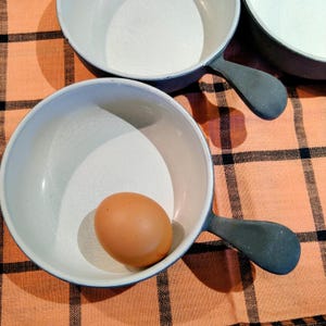 May include: Three white enamel pans with gray handles are arranged on an orange and black checkered surface. One pan contains a brown egg. The pans have a white interior and a blue rim.