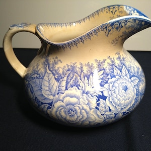 May include: Cream-colored pitcher with a blue floral design. The pitcher features a bulbous body, a curved handle, and a spout. The blue floral pattern includes large flowers and foliage. The rim and handle have blue decorative accents.