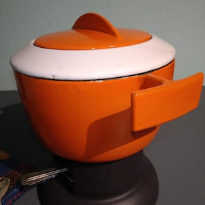 May include: An orange and white enamelware cooking pot with a lid and handle. The pot has a rounded shape with a white band around the top and a small orange handle. The lid has a fin-shaped handle. A vintage kitchenware item.