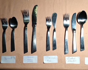 May include: A collection of stainless steel flatware arranged in pairs and singles, including a small spoon, forks, knives, and large spoons. Each set is labeled with white paper tags reading Café, Dessert, Viande, Poisson, Entrée, and Soupe.