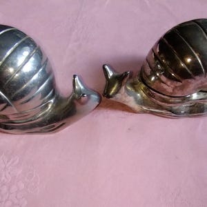 Könnte beinhalten: Zwei silberfarbene Schneckenfiguren. Die Schnecken haben detaillierte Schalenmuster und sind einander zugewandt. Die Figuren befinden sich auf einer rosa Oberfläche, was auf einen dekorativen oder sammelbaren Gegenstand hindeutet.