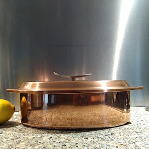 May include: A copper-colored cooking pot with a lid. The pot has a wide, shallow shape with a flat rim and a curved handle on the lid. The pot is sitting on a speckled countertop, with a lemon on the left side.