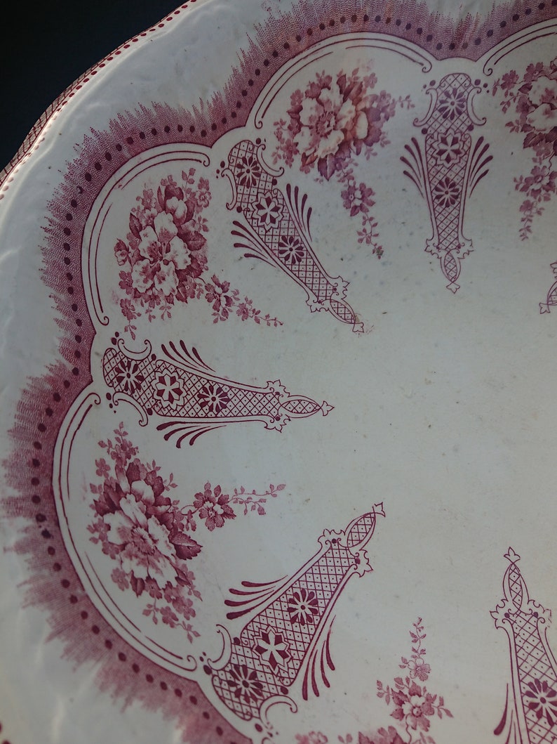 May include: A white plate with a scalloped edge and a burgundy floral and geometric pattern. The plate features a repeating design of flowers and decorative shapes, creating a vintage aesthetic.