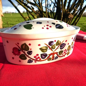 May include: A white ceramic casserole dish with a lid, decorated with a floral pattern of red berries, green and purple leaves, and brown branches. The dish is rectangular and sits on a red tablecloth.