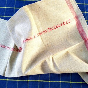 Peut inclure: Un tissu de lin plié, de couleur crème, avec des rayures et du texte rouges. Le texte rouge indique "PAPIERS A CIGARETTES ZIG-ZAG & O.C.B.". Le tissu est posé sur une surface à carreaux bleue et jaune.