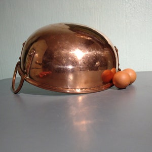 May include: A copper bowl with a handle on the side. The bowl is sitting on a grey surface with three brown eggs next to it.