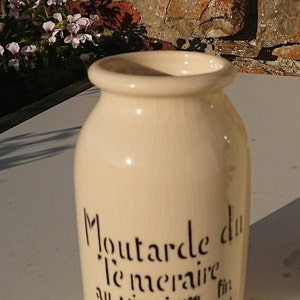 Puede incluir: Tarro de cer&aacute;mica color crema con texto negro "Moutarde du T&eacute;m&eacute;raire au vinaigre fin Dijon BORNIER." El tarro tiene forma redondeada y una abertura ancha. El fondo incluye una pared de ladrillo y flores.