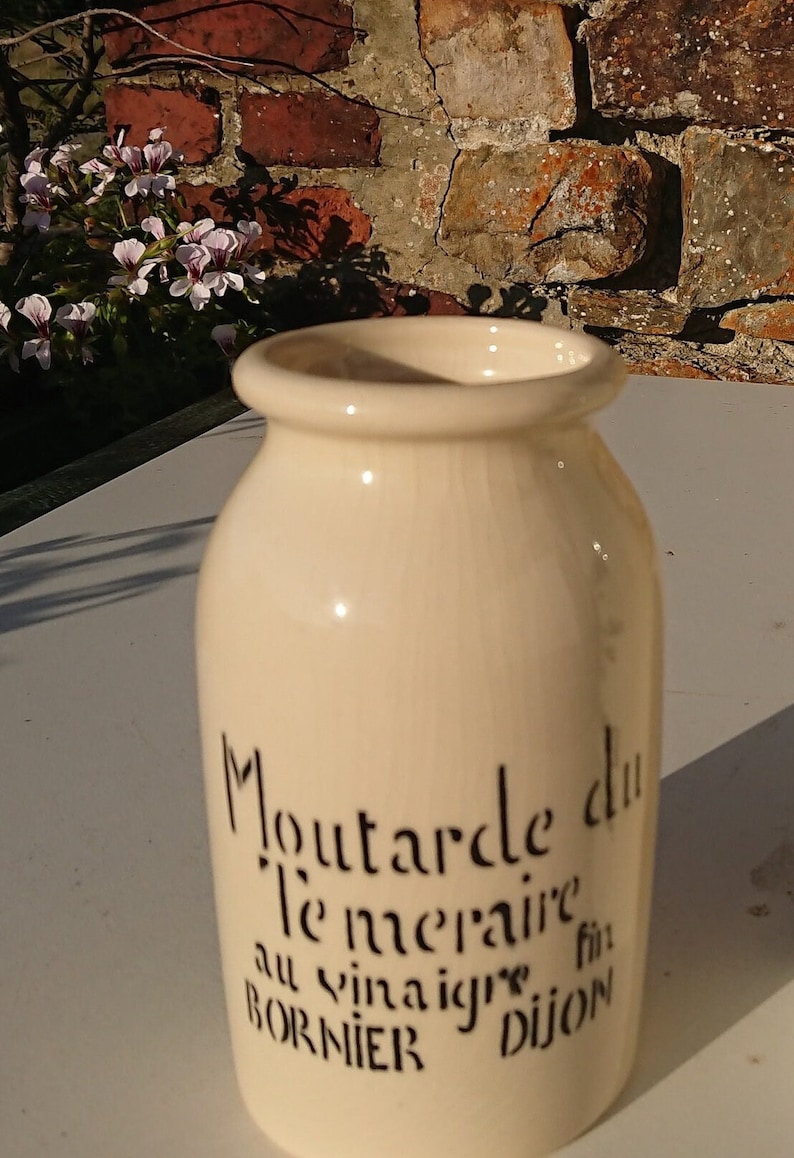 Puede incluir: Tarro de cer&aacute;mica color crema con texto negro "Moutarde du T&eacute;m&eacute;raire au vinaigre fin Dijon BORNIER." El tarro tiene forma redondeada y una abertura ancha. El fondo incluye una pared de ladrillo y flores.