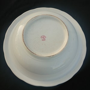 May include: A white ceramic bowl with a scalloped edge, viewed from the underside. The bowl has a slightly textured surface with visible crackling. A red stamp with text is visible in the center.