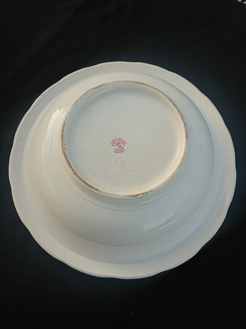 May include: A white ceramic bowl with a scalloped edge, viewed from the underside. The bowl has a slightly textured surface with visible crackling. A red stamp with text is visible in the center.