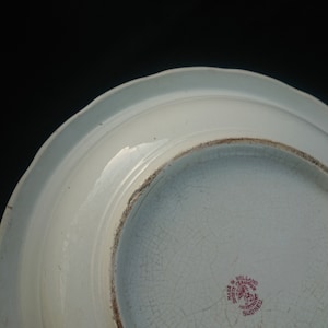 May include: A white ceramic plate with a scalloped edge, viewed from the underside. The plate has a cracked, aged appearance. A red stamp reads "Made in Holland" and "Societe Ceramique Maastricht Sidney".