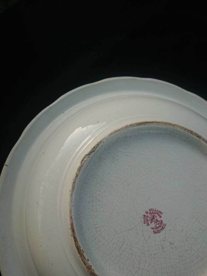 May include: A white ceramic plate with a scalloped edge, viewed from the underside. The plate has a cracked, aged appearance. A red stamp reads "Made in Holland" and "Societe Ceramique Maastricht Sidney".