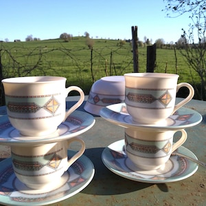 Puede incluir: Cuatro tazas y platillos color crema apilados sobre una mesa. Cada taza y platillo presenta un diseño geométrico en tonos verdes, melocotón y azul. Una cuchara descansa sobre la mesa. El fondo muestra un campo verde y cielo azul.