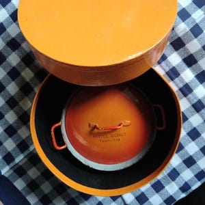 May include: An orange, round container with a matching lid. Inside, a smaller orange pot with red handles and the text "HOTEL DENET TEMPLOUA" is visible. The container sits on a blue and white checkered surface.