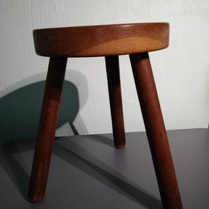 May include: A wooden stool with a round seat and three legs. The stool is made of dark brown wood and has a natural finish.