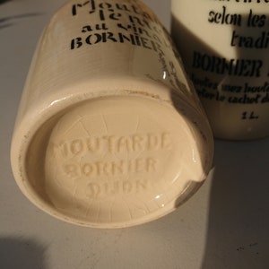 Puede incluir: Dos tarros de mostaza de cer&aacute;mica blanquecina con texto negro. Uno est&aacute; tumbado, mostrando la base con la marca "Moutarde Bornier Dijon". El otro est&aacute; de pie, con el texto "vieilli en foudre selon les traditions."