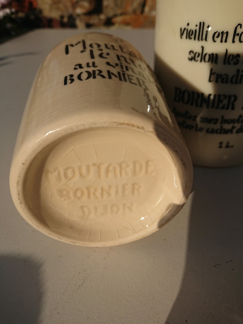 Puede incluir: Dos tarros de mostaza de cer&aacute;mica blanquecina con texto negro. Uno est&aacute; tumbado, mostrando la base con la marca "Moutarde Bornier Dijon". El otro est&aacute; de pie, con el texto "vieilli en foudre selon les traditions."