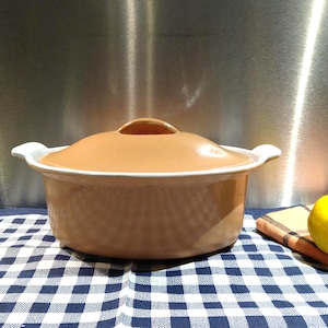 May include: A tan ceramic casserole dish with a matching lid and white handles. The dish sits on a blue and white checkered tablecloth. A lemon and a yellow and orange checkered napkin are in the background.