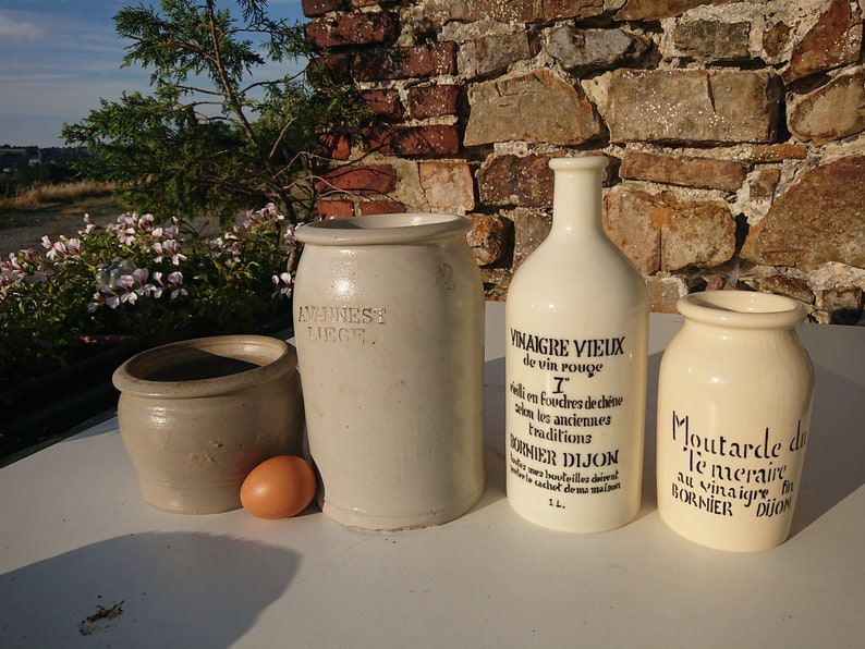 Puede incluir: Un surtido de recipientes de cer&aacute;mica vintage y una botella, con texto en franc&eacute;s, sobre una superficie blanca. Los recipientes son de tonos beige y crema, con una botella etiquetada "Vinaigre Vieux". Un huevo descansa cerca de los recipientes.
