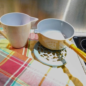May include: A cream-colored pitcher and a small yellow saucepan with a wooden handle are displayed on a colorful plaid tablecloth. The pitcher has a light blue interior, and the saucepan sits on a decorative trivet.