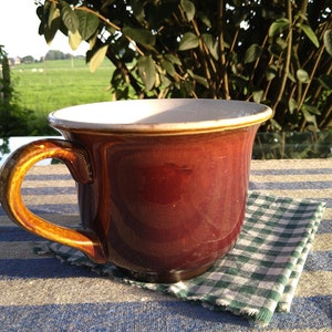 Könnte beinhalten: Eine große, braune Keramiktasse mit breitem Rand und gebogenem Henkel. Die Tasse steht auf einer blau-weiß gestreiften Tischdecke, neben einer grün-weiß karierten Serviette. Der Hintergrund zeigt Grün und eine sonnige Szene im Freien.
