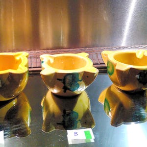 Op de afbeelding: Drie keramische asbakken met een gele binnenkant en een bruine buitenkant. Elke asbak heeft een unieke vorm met een uitlopende rand en een kleine rustplaats voor sigaretten. De asbakken worden op een reflecterend oppervlak weergegeven.