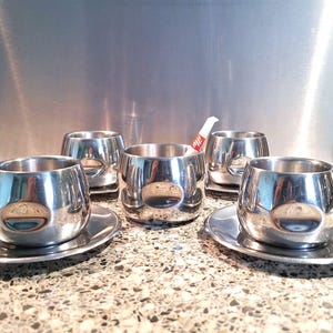 Könnte beinhalten: Ein Set aus vier polierten Edelstahl-Espressotassen mit passenden Untertassen. Die Tassen haben ein abgerundetes, doppelwandiges Design mit reflektierender Oberfläche. Eine Tasse enthält ein kleines weißes Tütchen mit rotem illy-Schriftzug.