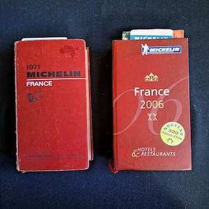 Puede incluir: Dos guías Michelin rojas de Francia. El libro de la izquierda está fechado en 1971 y presenta el logotipo y texto de Michelin. El libro de la derecha es de 2006, titulado France Hotels & Restaurants, e incluye una pegatina circular Nouveau 320 maisons d'hôte.