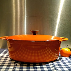 May include: An orange enameled cast iron Dutch oven with a lid and side handles. The pot sits on a blue and white checkered cloth. A small orange fruit with a green stem is next to the pot.