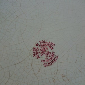 May include: Close-up of a white ceramic plate with a cracked glaze pattern. A red circular stamp reads "MADE IN HOLLAND CERAMIQUE SOCIETE MAESTRICHT SUD NEIJ."