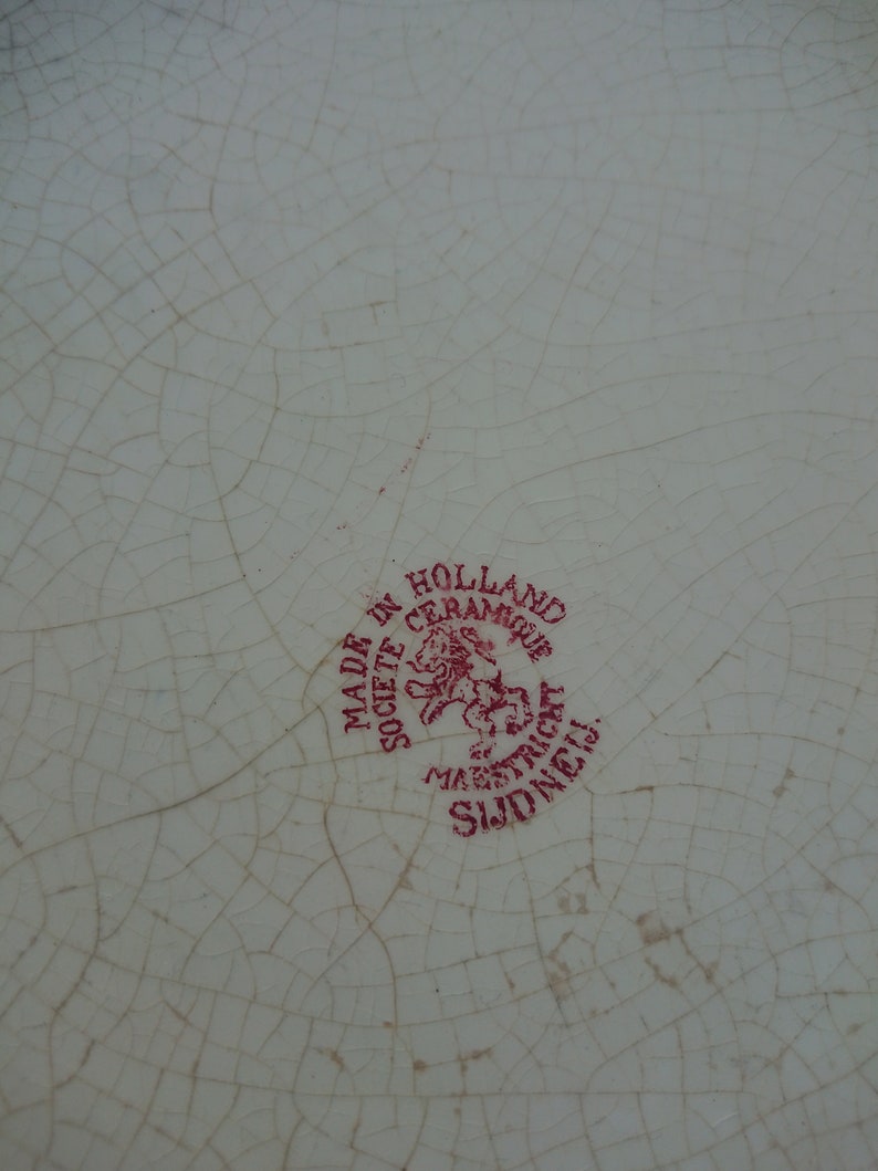 May include: Close-up of a white ceramic plate with a cracked glaze pattern. A red circular stamp reads "MADE IN HOLLAND CERAMIQUE SOCIETE MAESTRICHT SUD NEIJ."