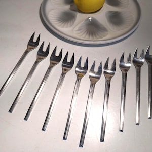 May include: A set of ten silver-toned cocktail forks with two prongs each. The forks are arranged in a fan shape on a white surface.