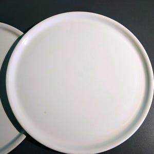 May include: Two white ceramic plates, one stacked on top of the other. The plates are round and have a slightly raised rim.