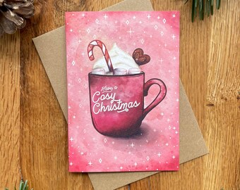 May include: A pink Christmas card with a red mug filled with hot chocolate, topped with whipped cream, a candy cane, and marshmallows. The text on the card reads "Have a Cosy Christmas".