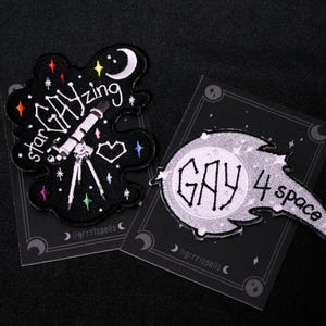 Op de afbeelding: Twee geborduurde patches op een zwart oppervlak. Eén patch is zwart met de woorden "star GAYzing" en een telescoopontwerp. De andere patch is wit met de woorden "GAY 4 space".