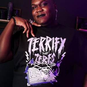 Peut inclure: T-shirt noir avec l'inscription "TERRIFY THE TERFS" dans une police gothique dégoulinante. En dessous, un livre ouvert avec l'inscription "TRANS RIGHTS ARE HUMAN RIGHTS" et un pentagramme. Le t-shirt est porté par une personne.