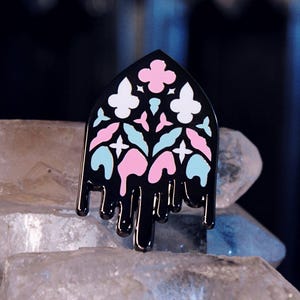 May include: Black enamel pin with a stained glass design. The pin features a black outline with pink, white, and light blue floral and geometric shapes. The bottom of the pin has a dripping effect. The pin is displayed on a bed of crystals.