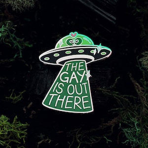 Pode incluir: Pin de esmalte verde com um alienígena de desenho animado em um disco voador. O texto diz "The Gay Is Out There".