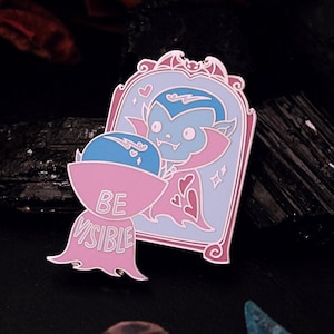 May include: A pink and blue enamel pin featuring a cartoon vampire looking into a mirror with the text "BE VISIBLE" on the bottom.