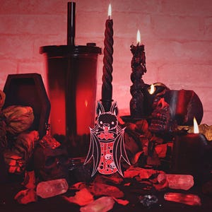 May include: A black and white bat-shaped enamel pin with a red and white boba tea cup in its mouth. The pin is on a dark red surface with black candles and other decorations.