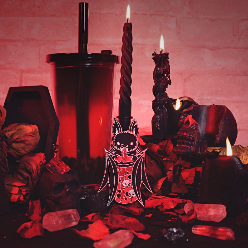 May include: A black and white bat-shaped enamel pin with a red and white boba tea cup in its mouth. The pin is on a dark red surface with black candles and other decorations.